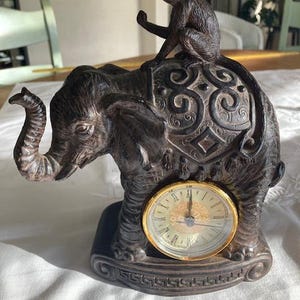 Puede incluir: Una figura esculpida de color marrón oscuro que representa un elefante con un mono posado en su lomo. El elefante presenta una silla de montar ornamentada con volutas y un reloj analógico redondo con borde dorado y números romanos incrustado.