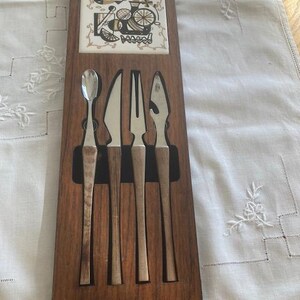 Op de afbeelding: Een rechthoekige houten wand-barset met een vierkante keramische tegel met een zwart-gouden grafische print van citrusslices en cocktailglazen. Vier roestvrijstalen bar-tools met houten handvatten zitten in een zwarte inzetstuk.