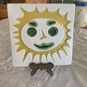 May include: A square white ceramic tile featuring a raised, yellow sun design with a green face. The sun has a smiling mouth, two circular green eyes, and a pointed nose. The tile is supported by a dark, ornate metal display stand.