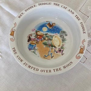 以下が含まれることがあります： 「Hey Diddle Diddle」の童謡のキャラクターが描かれた白い陶器製のボウル。月を飛び越える牛、バイオリンを弾く猫、スプーンと皿のカラフルなイラストが中央にあり、縁には「HEY, DIDDLE, DIDDLE! THE CAT AND THE FIDDLE THE COW JUMPED OVER THE MOON」という文字が記されています。