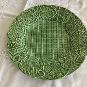 Może przedstawiać: Okrągły ceramiczny talerz w kolorze szałwiowej zieleni z centralnym wzorem kratki przypominającym pleciony koszyk. Brzeg talerza ozdobiony jest serią wypukłych, szczegółowych motywów liści ułożonych w okrągły, zachodzący na siebie wzór.