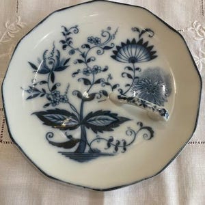 May include: White ceramic plate with a blue floral design and a built-in spoon rest. The plate has an octagonal shape with a dark blue trim. The floral pattern features stylized flowers and leaves in shades of blue.