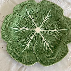 Puede incluir: Un plato de cerámica redondo con forma de hoja de col verde. La superficie presenta un diseño texturizado y en relieve que imita las venas y los bordes ondulados de una hoja vegetal, con un punto central blanco donde convergen las venas.