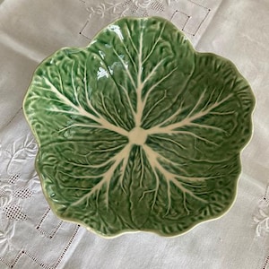 May include: A ceramic bowl shaped like a cabbage leaf with a glossy finish. The bowl features a vibrant green colour with prominent white veins radiating from the centre, mimicking the natural texture and appearance of a fresh cabbage leaf.