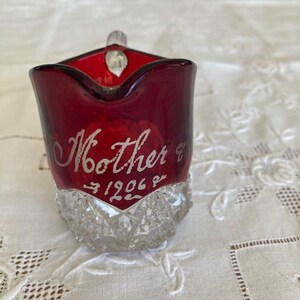 Può includere: Una piccola lattiera in vetro rosso rubino con una base trasparente a motivo diamantato. La parte superiore rossa liscia è incisa con il testo Mother e l'anno 1906. La lattiera presenta un manico curvo e un beccuccio per versare.