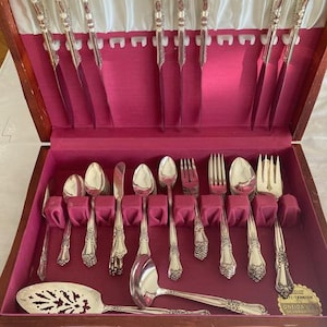 Op de afbeelding: Een houten bestekdoos met een magenta interieur met een set verzilverd bestek. De set bevat messen, lepels, vorken, een soeplepel en een taartschep. Een gouden label in de hoek vermeldt de tekst Anti-Tarnish Oneida Ltd.