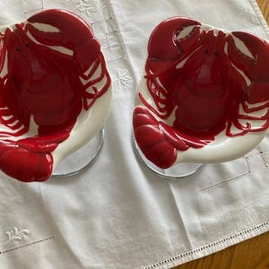 May include: Two matching ceramic bowls featuring a glossy red lobster relief design centered on a white background. Each bowl has a rounded shape with the lobster's claws and tail extending toward the edges, creating a dimensional appearance.