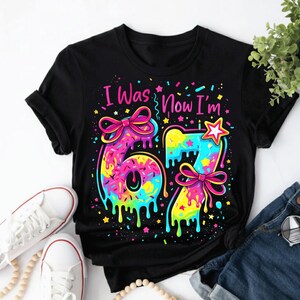 May include: Black t-shirt with the text "I Was Now I'm" above a colorful "67" design. The numbers are decorated with sprinkles, bows, and dripping paint effects, with a star on the "7".
