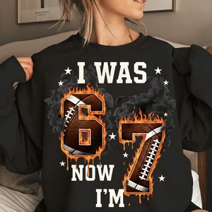 Pode incluir: Camisola preta com o texto branco "I WAS 67 NOW I'M". Os n&uacute;meros 6 e 7 s&atilde;o projetados como bolas de futebol com chamas. Estrelas brancas espalhadas ao redor do texto.