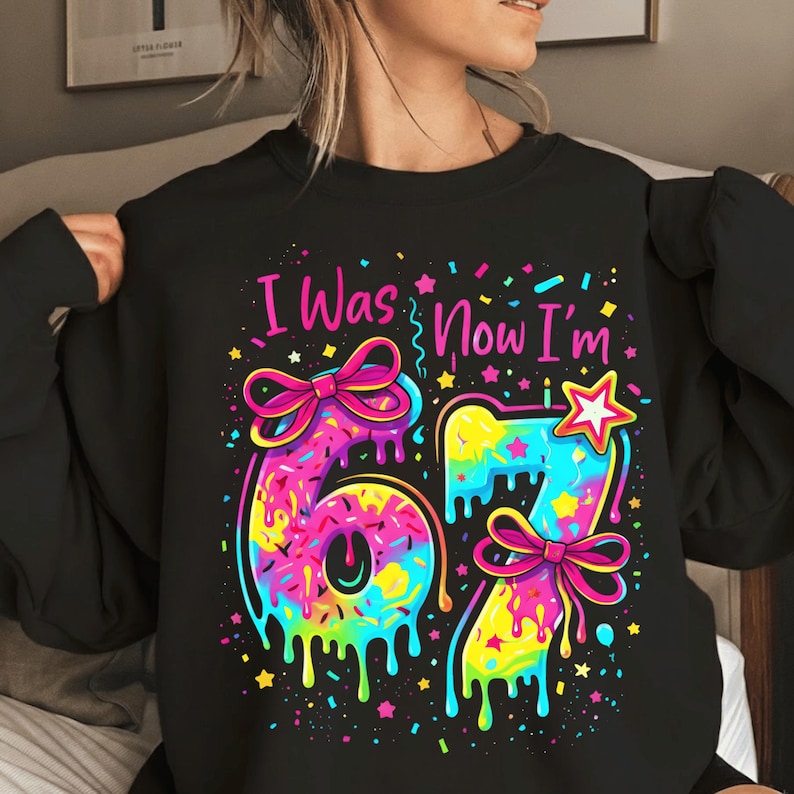 May include: Black sweatshirt with a colorful graphic that reads "I Was Now I'm 67". The numbers are designed to look like donuts with dripping icing and bows. The design includes stars and confetti.