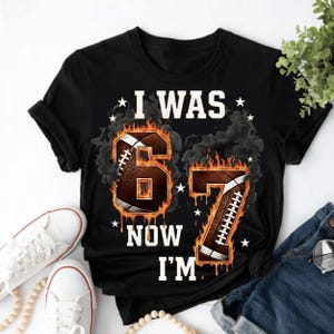 Pode incluir: Camiseta preta com o texto branco "I WAS 67 NOW I'M". Os n&uacute;meros 6 e 7 s&atilde;o projetados como bolas de futebol americano com efeitos de chamas e fuma&ccedil;a. Estrelas brancas espalhadas.