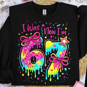 May include: Black sweatshirt with the text "I Was Now I'm" above the number 67. The numbers are colorful with a dripping effect, bows, and a star. The design is in bright pink, yellow, blue, and green.