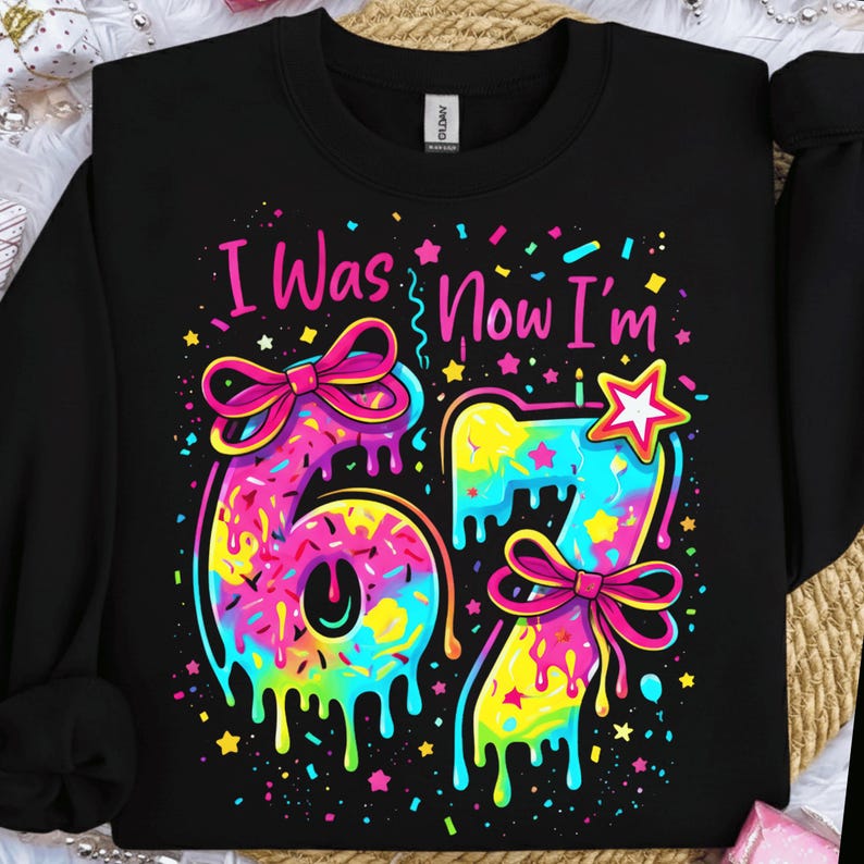 May include: Black sweatshirt with the text "I Was Now I'm" above the number 67. The numbers are colorful with a dripping effect, bows, and a star. The design is in bright pink, yellow, blue, and green.