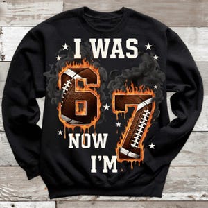 Pode incluir: Camisola preta com o texto "I WAS 67 NOW I'M" em branco. Os n&uacute;meros 6 e 7 s&atilde;o projetados como bolas de futebol americano em chamas, com estrelas brancas e detalhes de fuma&ccedil;a. Uma pe&ccedil;a de vestu&aacute;rio com tema desportivo.