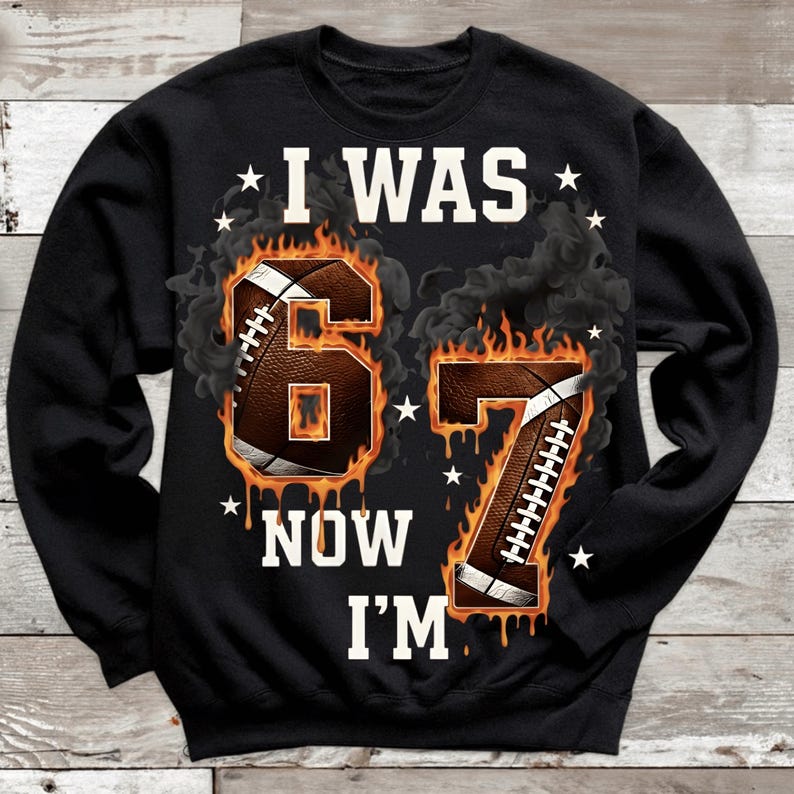 Pode incluir: Camisola preta com o texto "I WAS 67 NOW I'M" em branco. Os n&uacute;meros 6 e 7 s&atilde;o projetados como bolas de futebol americano em chamas, com estrelas brancas e detalhes de fuma&ccedil;a. Uma pe&ccedil;a de vestu&aacute;rio com tema desportivo.