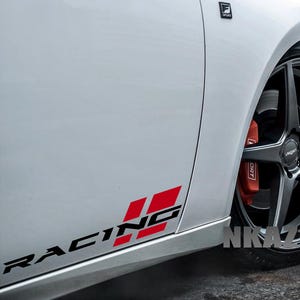Pode incluir: Carro branco com um gráfico "RACING" preto e vermelho na lateral. O carro tem uma roda preta com uma pinça de freio vermelha. A imagem também mostra o texto "NKAZ1080".