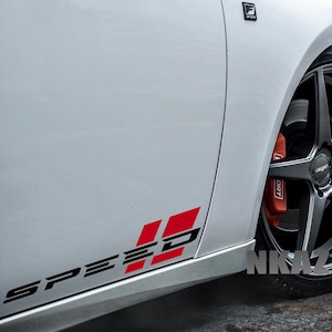 May include: White car with black rims and red brake calipers. The word "SPEED" is written in black with red accents on the side of the car. The car has a sporty, modern design.