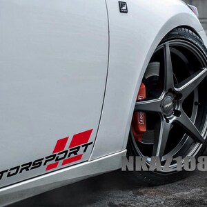 Op de afbeelding: Witte auto met een zwart en rode "Motorsport" sticker op de zijkant. De auto heeft een zwarte velg met een vijfspaaks ontwerp en een rode remklauw. De tekst "NKAZ1080" is zichtbaar bij het wiel.
