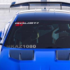 Puede incluir: Un coche deportivo azul con un alerón negro y una pancarta en el parabrisas que dice "ROUSH" en blanco y rojo. El texto "NKAZ1080" es visible en la parte inferior del parabrisas.