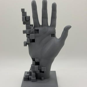 Pode incluir: Uma escultura cinza impressa em 3D de uma mão, com um efeito pixelizado no pulso e antebraço. A mão está aberta, com os dedos estendidos. A escultura é montada sobre uma base quadrada.