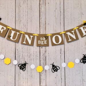 Puede incluir: Una pancarta marr&oacute;n y blanca con el texto "Fun to Bee One" y decoraciones de abejas amarillas y negras. La pancarta est&aacute; atada con una cinta amarilla y lazos negros. Una guirnalda separada de abejas negras, c&iacute;rculos blancos y hex&aacute;gonos amarillos est&aacute; atada debajo de la pancarta.