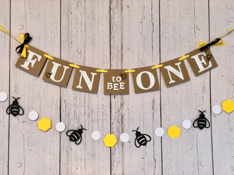 Puede incluir: Una pancarta marr&oacute;n y blanca con el texto "Fun to Bee One" y decoraciones de abejas amarillas y negras. La pancarta est&aacute; atada con una cinta amarilla y lazos negros. Una guirnalda separada de abejas negras, c&iacute;rculos blancos y hex&aacute;gonos amarillos est&aacute; atada debajo de la pancarta.