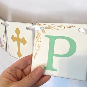 May include: A white banner with the word "Happy" spelled out in green letters. Each letter is on a separate square card with a gold cross on one card and gold decorative flourishes on the other cards. The banner is strung together with white ribbon.