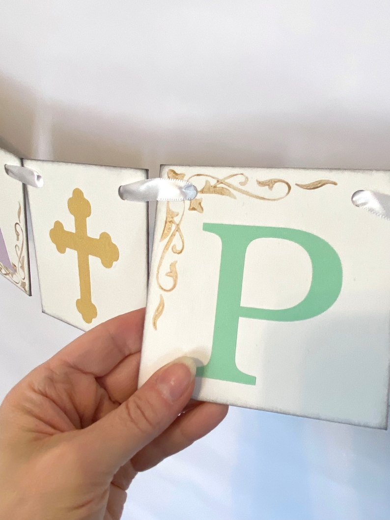 May include: A white banner with the word "Happy" spelled out in green letters. Each letter is on a separate square card with a gold cross on one card and gold decorative flourishes on the other cards. The banner is strung together with white ribbon.
