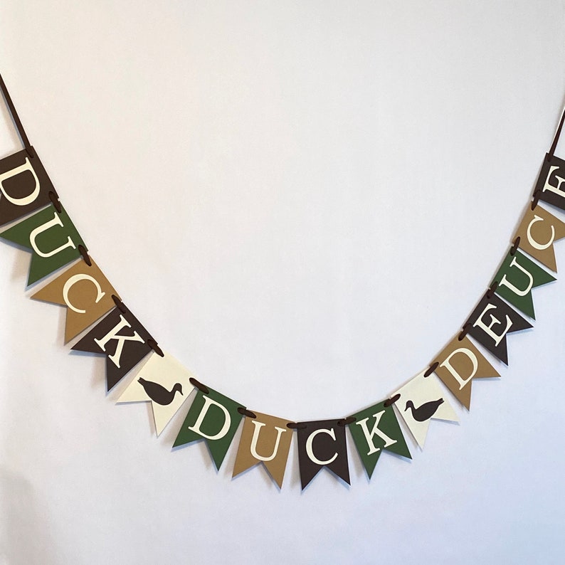 May include: A brown, green, and white banner with the words "Duck, Duck" in white letters. The banner is made of felt and has a string for hanging.