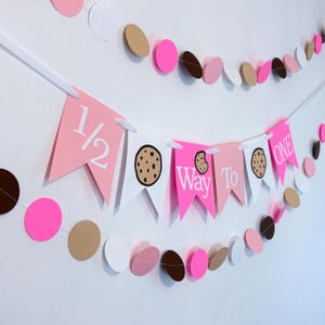 May include: A pink, white, and brown banner with the text "1/2 Way To One" and cookie graphics. The banner is strung with a white ribbon and is decorated with matching pink, brown, and white circles.