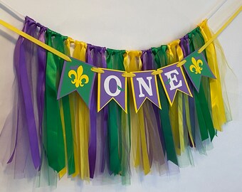 May include: Purple, green, and yellow tulle and ribbon banner with the word "ONE" in white letters on purple flags and fleur-de-lis on green flags.