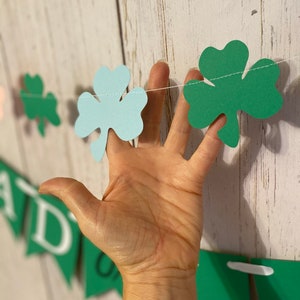 Pu&ograve; includere: Una ghirlanda di trifogli verde e blu con uno spago bianco. La ghirlanda &egrave; appesa a un muro di legno bianco.
