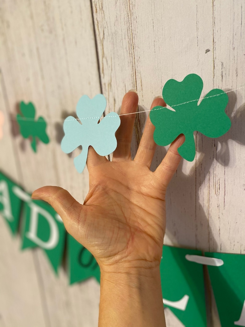 Pu&ograve; includere: Una ghirlanda di trifogli verde e blu con uno spago bianco. La ghirlanda &egrave; appesa a un muro di legno bianco.