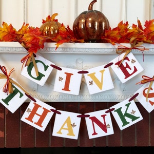 K&ouml;nnte beinhalten: Ein Thanksgiving-Banner mit den Worten "Give Thanks" in wei&szlig;en Buchstaben auf wei&szlig;em Hintergrund. Die Buchstaben sind mit Herbstbl&auml;ttern verziert und mit rotem Band zusammengebunden. Das Banner h&auml;ngt an einem wei&szlig;en Kaminsims mit Herbstbl&auml;ttern und einem K&uuml;rbis.