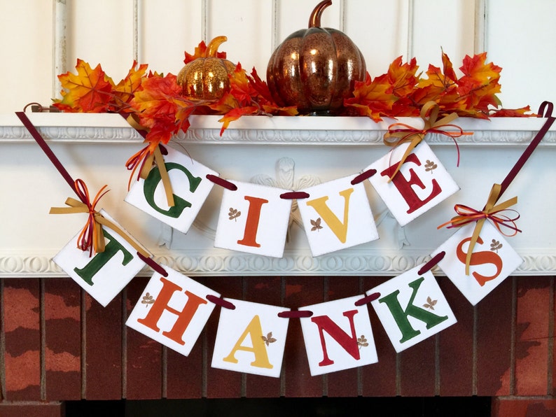K&ouml;nnte beinhalten: Ein Thanksgiving-Banner mit den Worten "Give Thanks" in wei&szlig;en Buchstaben auf wei&szlig;em Hintergrund. Die Buchstaben sind mit Herbstbl&auml;ttern verziert und mit rotem Band zusammengebunden. Das Banner h&auml;ngt an einem wei&szlig;en Kaminsims mit Herbstbl&auml;ttern und einem K&uuml;rbis.