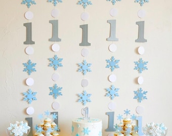 May include: A party display featuring a white cake topped with a number one candle, surrounded by cupcakes and snowflake-shaped cookies. The background consists of vertical garlands with light blue paper snowflakes, white circles, and the number one.