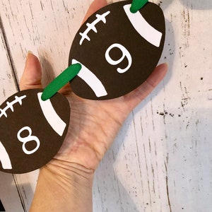 May include: A green and brown football-shaped banner with the numbers 8 and 9 printed on it. The banner is made of paper and has a white string running through it.