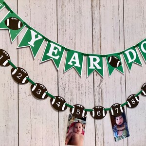 May include: Green and white banner with the text "1st Year Down" and a football design. A second banner with the text "YEAR" is strung below. A string of football-shaped decorations with numbers 1 through 10 are strung below the banners. Photos are attached to the footballs.