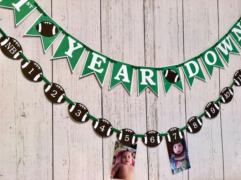 May include: Green and white banner with the text "1st Year Down" and a football design. A second banner with the text "YEAR" is strung below. A string of football-shaped decorations with numbers 1 through 10 are strung below the banners. Photos are attached to the footballs.