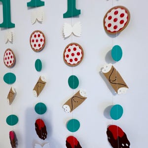 May include: A string of Italian-themed decorations, including pizza slices, cannoli, bow tie pasta, and green circles. The decorations are strung together with white string and are against a white background. The number "1" is also included in the design.