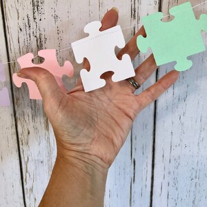 May include: A string of pastel colored paper puzzle pieces, strung together with white thread. The pieces are in shades of lavender, pink, white, and green.