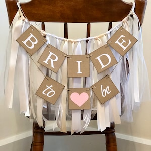 May include: A brown chair with a white and beige tulle and ribbon garland that says "Bride to be" with a pink heart in the middle.