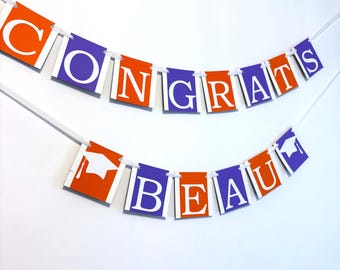 May include: A two-tiered banner featuring alternating orange and purple rectangular cardstock panels strung on white ribbon. The panels display the white text CONGRATS BEAU with two white graduation cap icons at the start and end of the phrase.