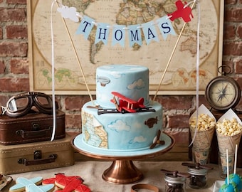 May include: A two-tier light blue cake decorated with clouds, a red biplane, and a map motif, topped with a banner reading THOMAS. The cake sits on a copper stand surrounded by airplane-shaped cookies, vintage suitcases, and popcorn.