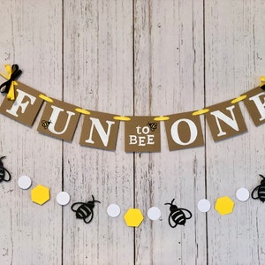 Puede incluir: Una pancarta marr&oacute;n y amarilla con el texto "Fun to Bee One" colgada sobre un fondo de madera blanca. Una hilera de hex&aacute;gonos amarillos y c&iacute;rculos blancos con recortes de abejas negras cuelga debajo de la pancarta.