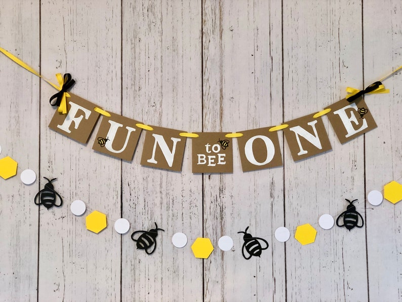 Puede incluir: Una pancarta marr&oacute;n y amarilla con el texto "Fun to Bee One" colgada sobre un fondo de madera blanca. Una hilera de hex&aacute;gonos amarillos y c&iacute;rculos blancos con recortes de abejas negras cuelga debajo de la pancarta.