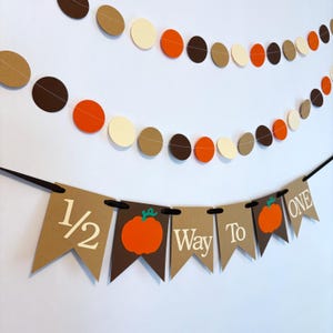 May include: A festive banner and garland set. The banner reads "1/2 Way To One" with orange pumpkin graphics. The garland features alternating brown, tan, orange, and cream-colored circles. Perfect for a half-birthday celebration.