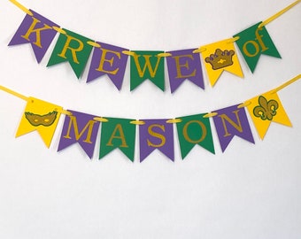 Könnte beinhalten: Ein Banner zum Thema Mardi Gras mit den Worten "Krewe of Mason" in goldenen Glitzerbuchstaben auf violetten und grünen Wimpeln. Die Wimpel haben goldene Glitzer-Mardi-Gras-Symbole, darunter eine Krone, eine Maske und eine Fleur-de-lis.