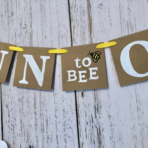 Puede incluir: Una pancarta de papel marr&oacute;n con letras blancas que deletrean "One to Bee" con una peque&ntilde;a ilustraci&oacute;n de abeja amarilla y negra a cada lado de la palabra "bee".