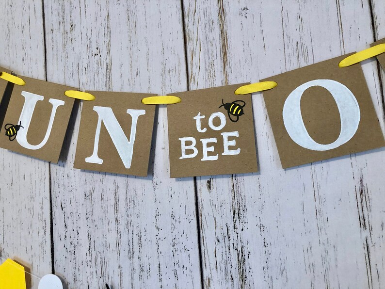 Puede incluir: Una pancarta de papel marr&oacute;n con letras blancas que deletrean "One to Bee" con una peque&ntilde;a ilustraci&oacute;n de abeja amarilla y negra a cada lado de la palabra "bee".